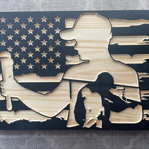 May include: Wooden wall art featuring a carved design of an American flag with a silhouette of a worker holding a wrench. The flag's stars and stripes are visible, with the worker's form in natural wood against a black background.