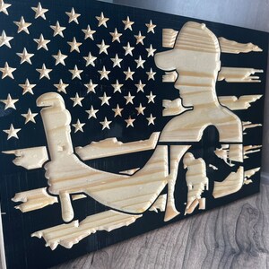 May include: Wooden American flag art piece featuring a carved silhouette of a worker holding a wrench. The flag design includes carved stars and stripes, with the worker's figure in natural wood against a black background. The artwork is displayed on a wooden surface.