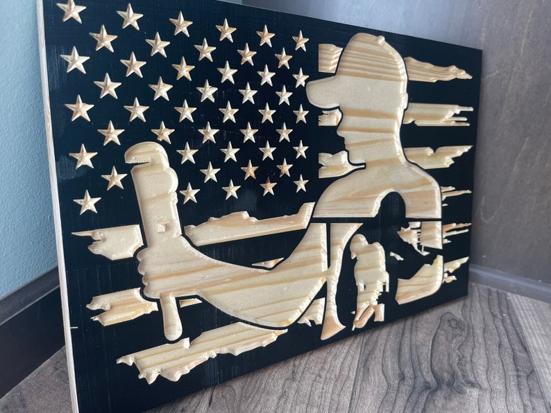 May include: Wooden American flag art piece featuring a carved silhouette of a worker holding a wrench. The flag design includes carved stars and stripes, with the worker's figure in natural wood against a black background. The artwork is displayed on a wooden surface.