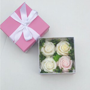 Puede incluir: Una caja de regalo rosa con una cinta y un lazo blancos, abierta para revelar una caja blanca con cuatro rosas de jab&oacute;n. Dos de las rosas son blancas y dos son rosas.