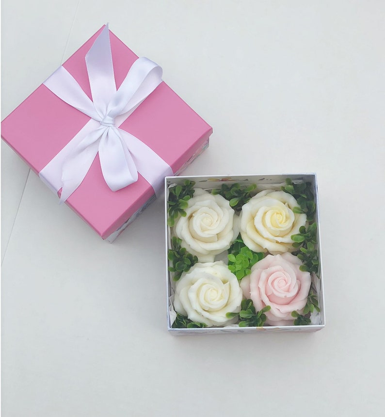 Puede incluir: Una caja de regalo rosa con una cinta y un lazo blancos, abierta para revelar una caja blanca con cuatro rosas de jab&oacute;n. Dos de las rosas son blancas y dos son rosas.