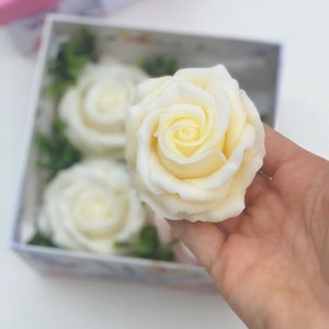 Puede incluir: Una caja de cuatro rosas de jab&oacute;n blanco. Las rosas est&aacute;n dispuestas en una caja con hojas verdes. Una rosa se sostiene en una mano.