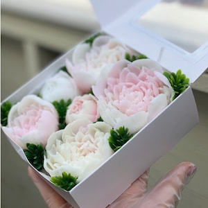 May include: A white box filled with pink and white peonies, arranged in a decorative pattern. The flowers are surrounded by green foliage.