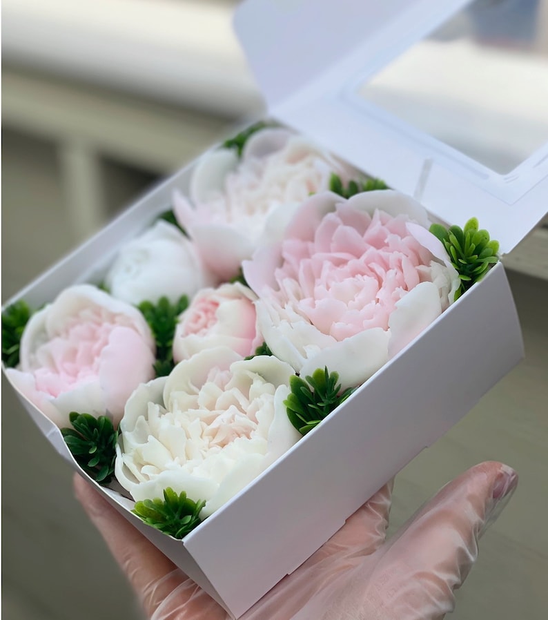May include: A white box filled with pink and white peonies, arranged in a decorative pattern. The flowers are surrounded by green foliage.