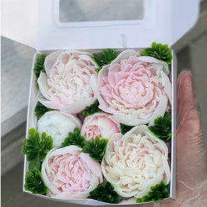 May include: A white gift box filled with six pink and white soap flowers. The flowers are arranged in a circular pattern and are surrounded by green leaves.
