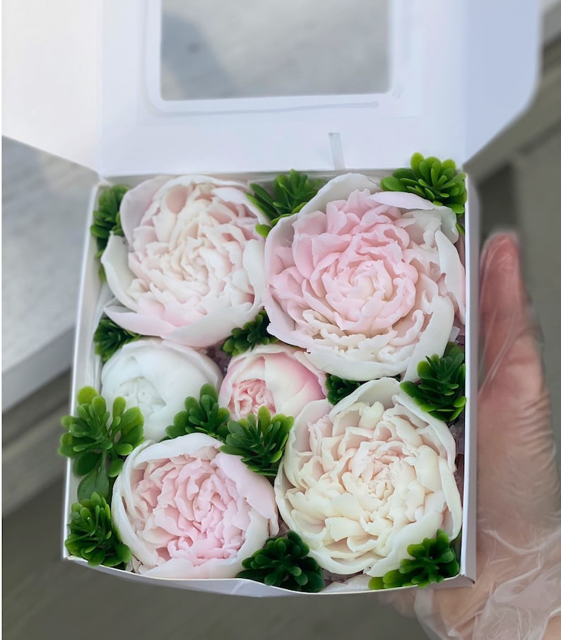 May include: A white gift box filled with six pink and white soap flowers. The flowers are arranged in a circular pattern and are surrounded by green leaves.