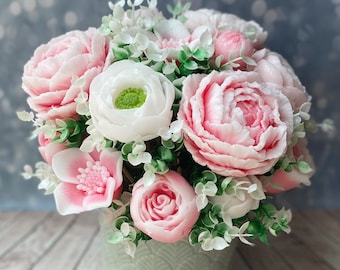 以下が含まれることがあります： ピンクと白の花のブーケ。シャクヤクやバラなど、薄緑色の花瓶に入っています。緑の葉が添えられ、背景はぼやけています。花は詳細でリアルな外観をしています。