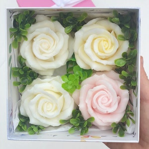 Puede incluir: Una caja de regalo blanca con cuatro rosas de jab&oacute;n dentro. Dos de las rosas son blancas y dos son rosas. Las rosas est&aacute;n rodeadas de hojas verdes.