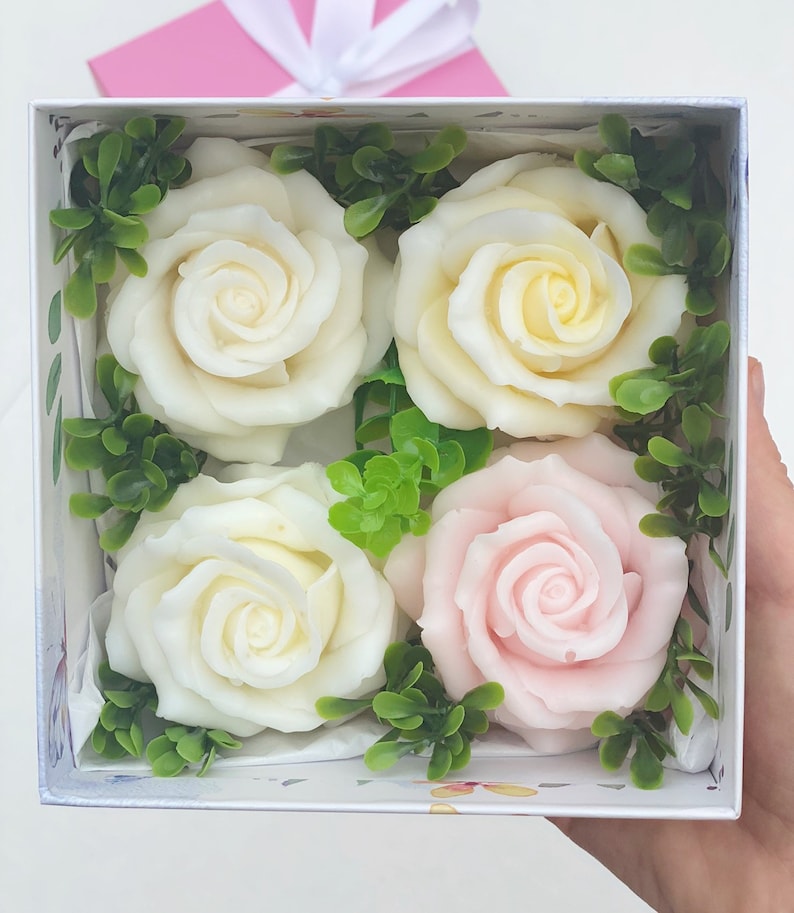 Puede incluir: Una caja de regalo blanca con cuatro rosas de jab&oacute;n dentro. Dos de las rosas son blancas y dos son rosas. Las rosas est&aacute;n rodeadas de hojas verdes.