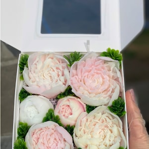 May include: A white box filled with six pink and white soap flowers. The flowers are arranged in a bouquet with green leaves. The box is open and the flowers are visible.