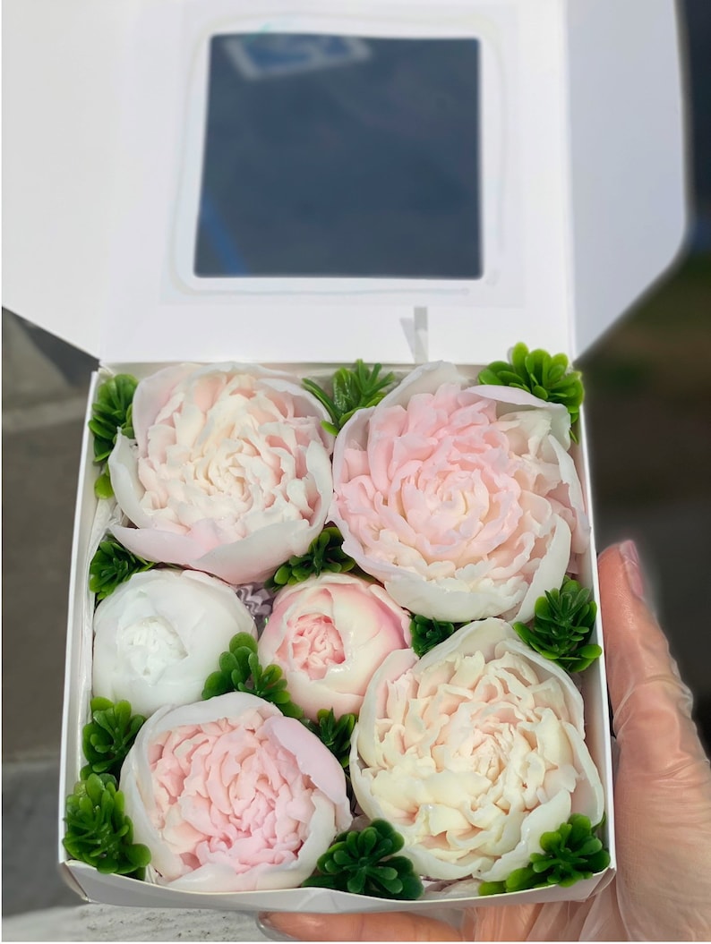 May include: A white box filled with six pink and white soap flowers. The flowers are arranged in a bouquet with green leaves. The box is open and the flowers are visible.