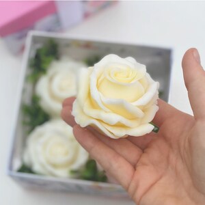 Puede incluir: Una caja de cuatro rosas de jab&oacute;n blanco, una de las cuales se sostiene en una mano. Las rosas est&aacute;n dispuestas en una caja con follaje verde.