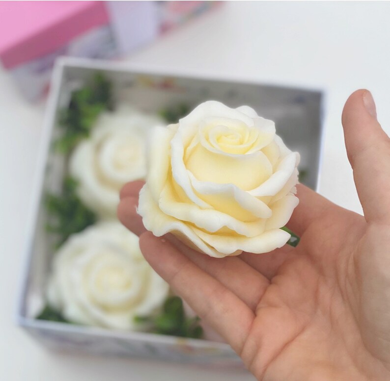 Puede incluir: Una caja de cuatro rosas de jab&oacute;n blanco, una de las cuales se sostiene en una mano. Las rosas est&aacute;n dispuestas en una caja con follaje verde.