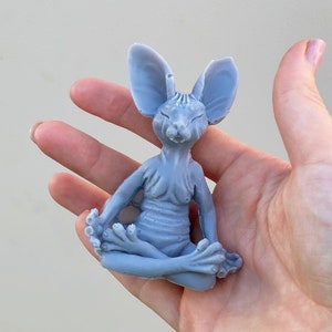 May include: A light blue ceramic figurine of a cat in a yoga pose. The cat has large ears and is sitting with its legs crossed.