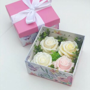 Puede incluir: Una caja de regalo rosa con una cinta blanca y un dise&ntilde;o floral. La caja est&aacute; abierta y contiene cuatro rosas de jab&oacute;n, dos blancas y dos de color crema, y una rosa de jab&oacute;n rosa.