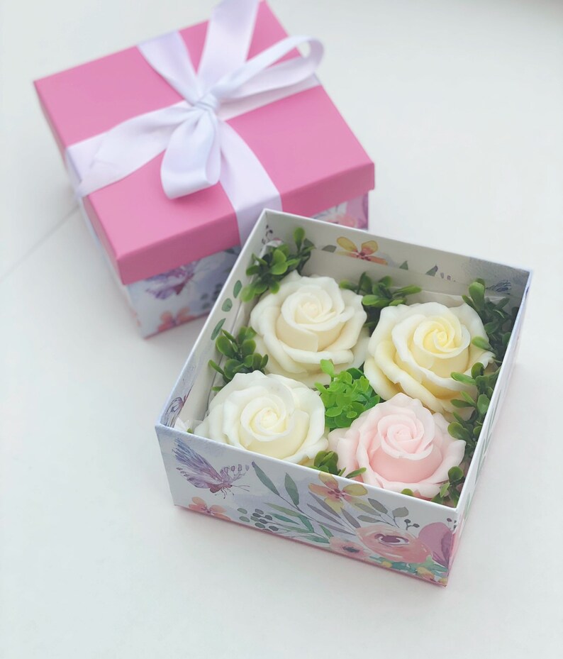 Puede incluir: Una caja de regalo rosa con una cinta blanca y un dise&ntilde;o floral. La caja est&aacute; abierta y contiene cuatro rosas de jab&oacute;n, dos blancas y dos de color crema, y una rosa de jab&oacute;n rosa.