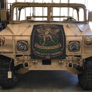 May include: A tan military Humvee, featuring a black banner with an illustration of St. Michael, the Guardian of the Fallen, and text. The vehicle has large tyres, headlights, and a protective front bumper. The image is taken from a front-facing perspective.