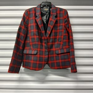 May include: A red and gray plaid blazer with a two-button closure and two front pockets. The blazer is hanging on a black hanger against a white background. The blazer has a classic, tailored design.