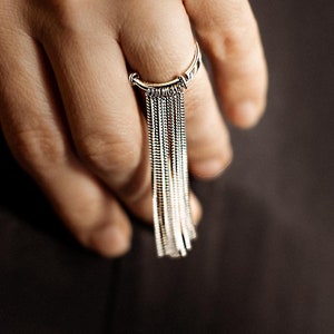 May include: A silver ring with a long chain tassel dangling from it. The ring is worn on a person's finger.