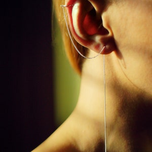 Puede incluir: Un pendiente de cadena de plata con una cadena larga y fina que cuelga del lóbulo de la oreja.