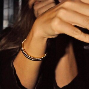 May include: A gold-colored bracelet with a simple, elegant design. The bracelet appears to be made of multiple bands, creating a layered effect. The jewelry is worn on the wrist, and the overall aesthetic is one of understated luxury.