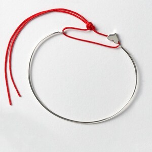 Könnte beinhalten: Ein silbernes Armreif-Armband mit einem kleinen Herz-Anhänger, der an einer roten Schnur befestigt ist. Die Schnur ist verknotet und hat lange Enden.