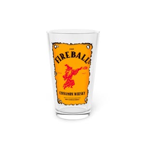 Clear pint glass centered on a rustic wooden table, printed with a Dungeons & Dragons parody “Fireball” label in orange and red reading “I cast FIREBALL!” with a wizard silhouette and “Cinnamon Whisky” text. White background.