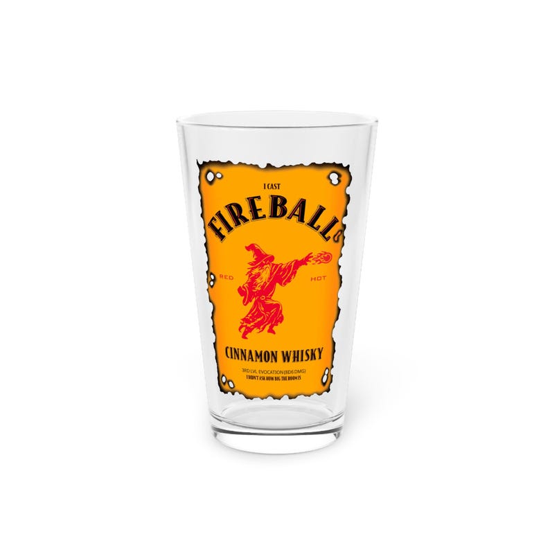 Clear pint glass centered on a rustic wooden table, printed with a Dungeons & Dragons parody “Fireball” label in orange and red reading “I cast FIREBALL!” with a wizard silhouette and “Cinnamon Whisky” text. White background.