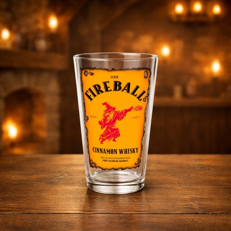 Clear pint glass centered on a rustic wooden table, printed with a Dungeons & Dragons parody “Fireball” label in orange and red reading “I cast FIREBALL!” with a wizard silhouette and “Cinnamon Whisky” text. Warm, blurred tavern background with candles and fireplace glow.