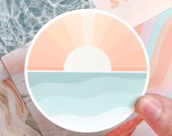 May include: A round sticker with a sunset over the ocean. The sunset is a peach and orange color with a white sun. The ocean is a light blue color with white waves.