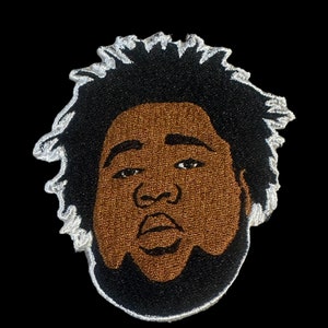 May include: Embroidered patch of a person's face with a black afro, brown skin, and a beard. The patch is outlined in white.