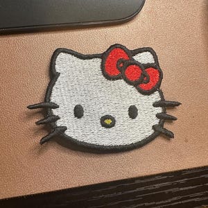May include: White embroidered Hello Kitty patch with a red bow. The patch has black outlines and a yellow nose.