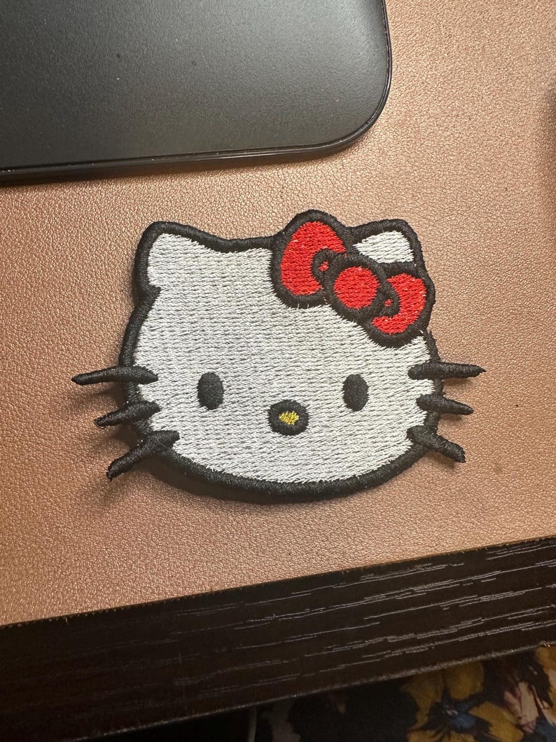 May include: White embroidered Hello Kitty patch with a red bow. The patch has black outlines and a yellow nose.