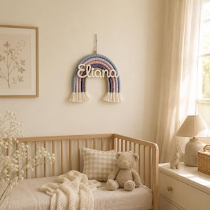 May include: A wall-hanging rainbow decoration made of wrapped yarn in shades of blue, purple, and cream, featuring the name Eliana in white cursive script across the center and finished with cream-colored fringe at both ends of the arch.