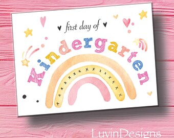 May include: A white card with a rainbow design and the text "✓ first day of kindergarten" in colorful letters. The rainbow is made of yellow, orange, pink, and blue watercolor. There are stars, hearts, and shooting stars scattered around the rainbow.