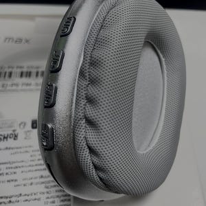 May include: A close-up of a grey over-ear headphone. The ear cup is covered in a grey mesh fabric. The side of the headphone has buttons with symbols. The headphone is resting on a white surface.