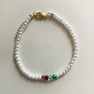 Könnte beinhalten: Ein weißes Perlenarmband mit einem goldenen Verschluss und einem kleinen Akzent aus roten, schwarzen und grünen Perlen.