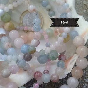 Peut inclure: Collier de perles aux couleurs pastel, rose, bleu, vert et blanc, avec des perles de différentes tailles et textures. Les colliers sont présentés à l'intérieur d'un grand coquillage. Le mot "Beryl" est écrit sur une bannière noire. Une pièce de monnaie est également visible.