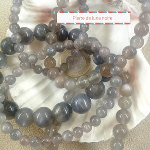 Op de afbeelding: Verschillende grijze maansteen kralenarmbanden van verschillende maten worden getoond op een witte zeeschelp. De kralen variëren van lichtgrijs tot donkergrijs. Een munt is zichtbaar in het midden. De tekst "Pierre de lune noire" staat op een wit en rood banner.