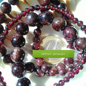 Peut inclure: Gros plan sur un collier de perles de pierres précieuses rouge foncé. Les perles sont rondes et ont une texture légèrement rugueuse. Les perles sont disposées en cercle, et une pièce de monnaie se trouve au centre du cercle. Le texte "GRENAT almandin" est écrit sur une étiquette verte.