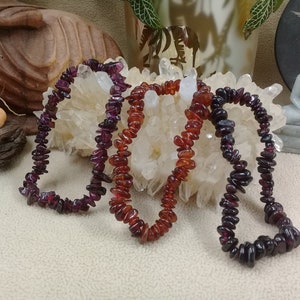 May include: Three gemstone chip bracelets in shades of red and purple. The bracelets are made of small, irregularly shaped stones strung together. The gemstones are displayed on a white surface with a natural background.