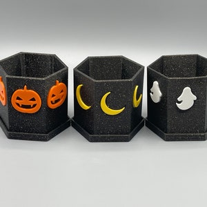 May include: Three black hexagonal planters with a speckled texture. The first features orange jack-o'-lantern faces, the second displays yellow crescent moons, and the third shows white ghost figures. Each container has a matching hexagonal base.