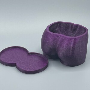 May include: A deep purple, glittery, hollow container shaped like a pair of buttocks, accompanied by a separate, matching figure-eight shaped flat tray. The object has a textured, matte finish and a wide, open top for storage or display purposes.