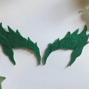 May include: Two symmetrical, jagged-edged mask pieces in a vibrant green glitter finish. Each piece features a pointed, flame-like silhouette with multiple sharp extensions, crafted from a textured material that reflects light across the surface.