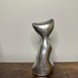 Puede incluir: Una escultura metálica de color plateado, lisa y con una forma fluida y abstracta. El objeto presenta una sección superior ancha y curva que se estrecha en un cuello fino, ensanchándose hacia la base, creando una superficie pulida y brillante.