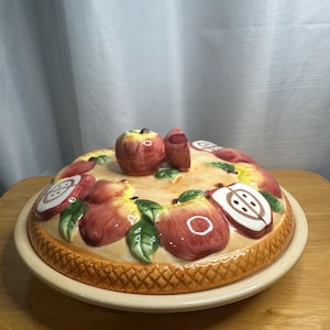 Puede incluir: Un molde redondo de cerámica para tartas con tapa a juego que presenta un motivo de manzanas en relieve pintado a mano. La tapa muestra manzanas rojas enteras, mitades de manzana y hojas verdes sobre un fondo crema, con un borde de corteza texturizado.
