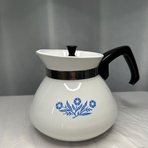 May include: A white teapot featuring a blue floral pattern on the front, a black handle, a black lid knob, and a metallic silver band encircling the upper portion of the body. The teapot has a rounded base and a wide opening with a flared rim.
