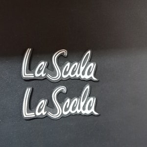 May include: Two silver-colored La Scala logos on a black background.