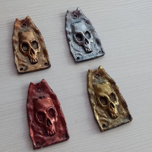 Puede incluir: Cuatro colgantes con forma de calavera en diferentes colores: oro, plata, cobre y bronce. Cada colgante está hecho de metal y tiene una superficie texturizada.