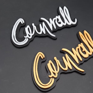 May include: Two versions of the word "Cornwall" in a stylized script font. One is white on black, the other is gold on black.